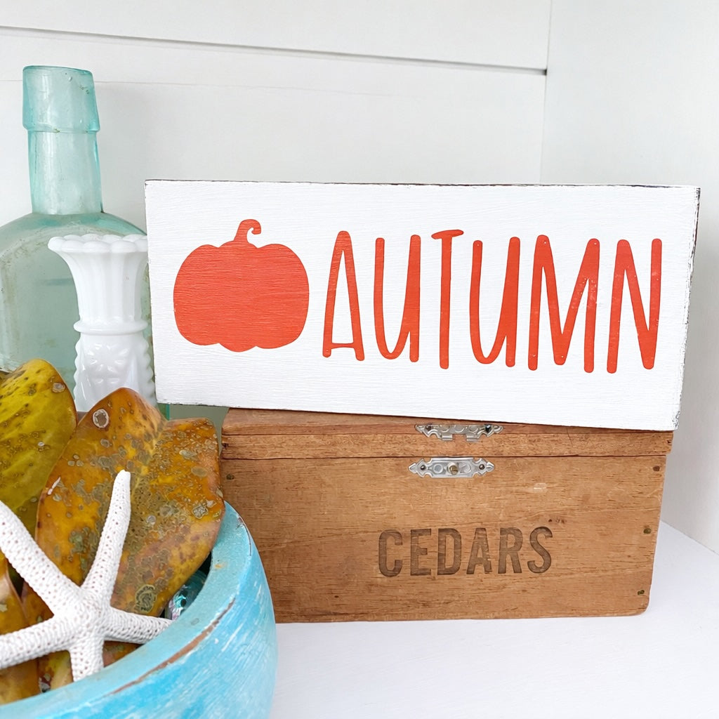 Wooden box with autumn-themed sign with pumpkin and 'AUTUMN' text, and decorative items on a white surface.