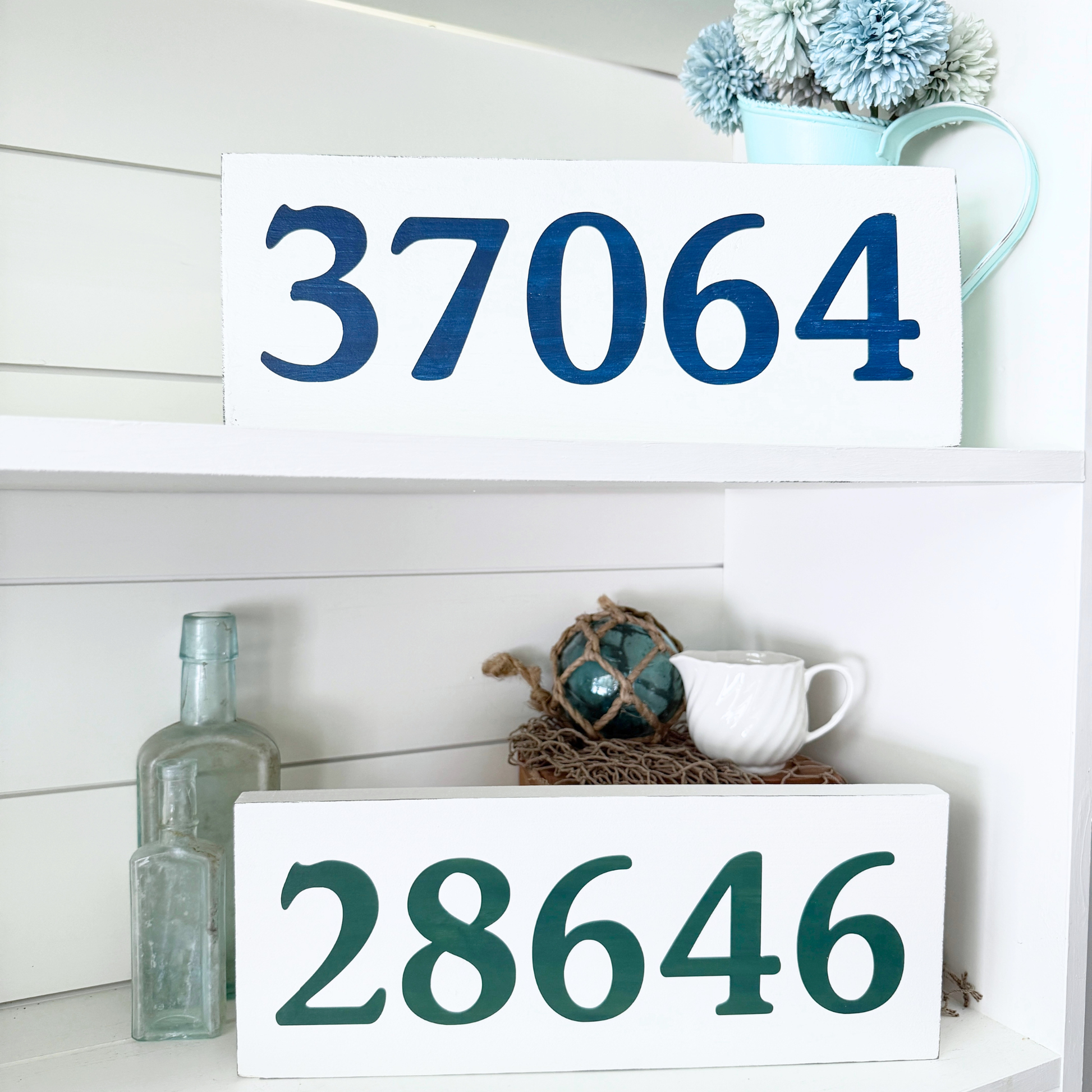 Two white wooden house numbers signs displayed on white shelves. The top sign reads '37064' in dark blue, and the bottom sign reads '28646' in dark green.