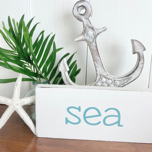 A decorative white wooden sign with the word 'sea' in laqua font, styled with a large distressed silver anchor, a white starfish, and green palm leaves on a wooden surface against a white paneled wall.