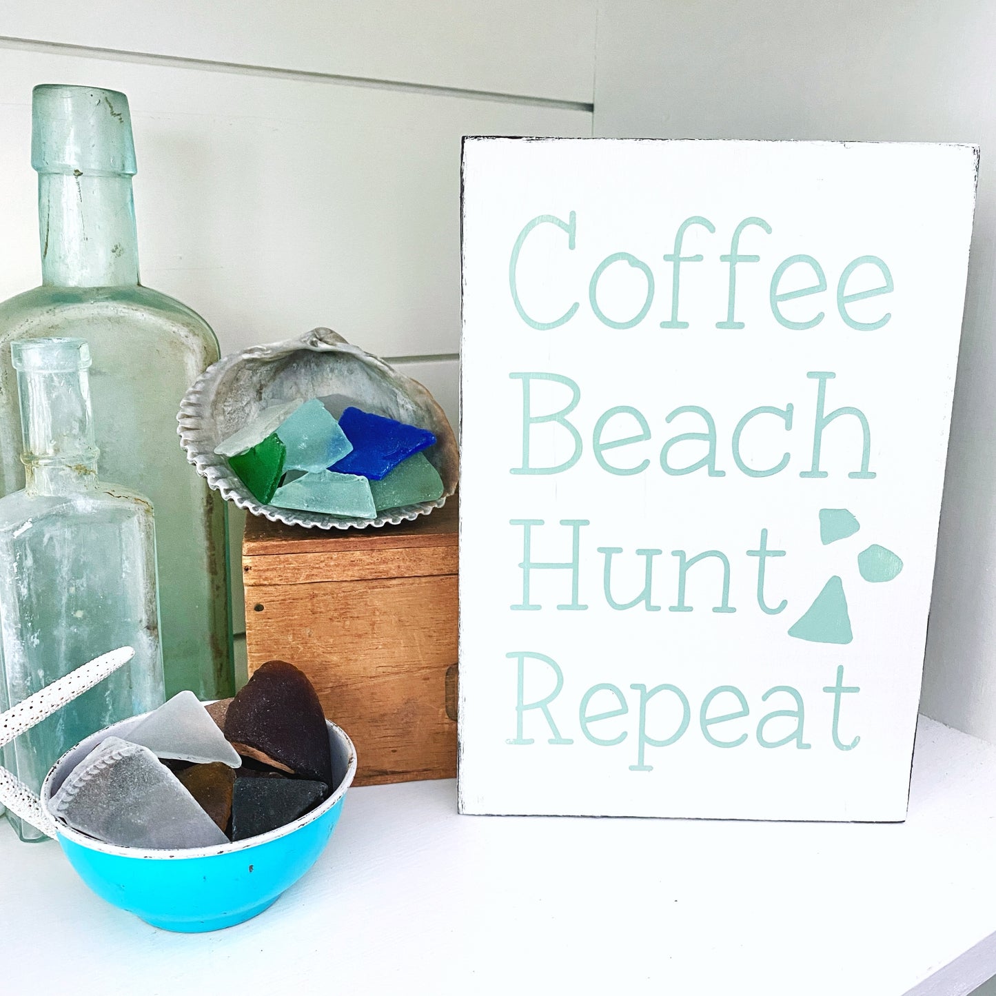Decorative setting with sea glass, bottles, and a sign reading 'Coffee Beach Hunt Repeat' on a white surface.