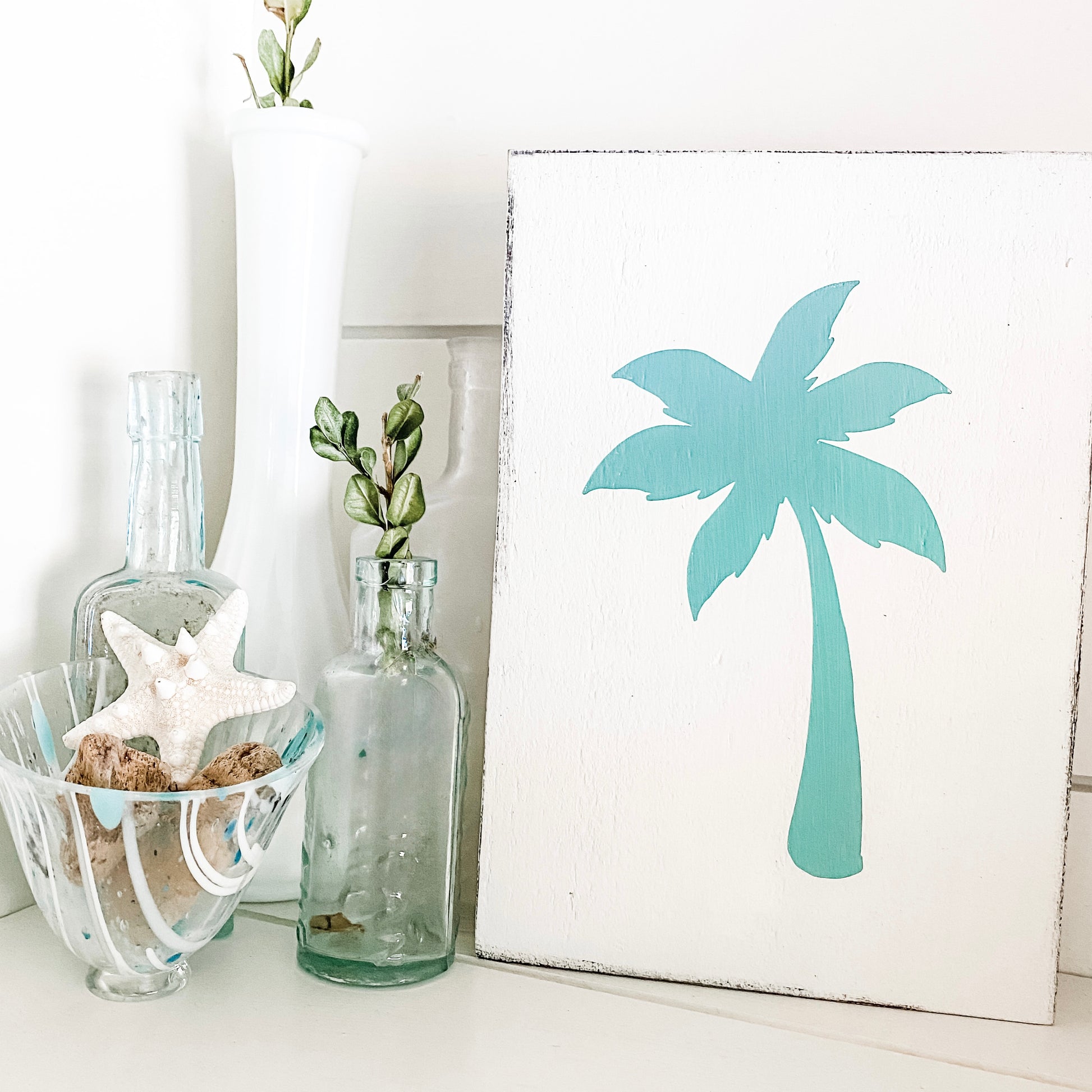 A white sign featuring a painted aqua palm tree silhouette, styled on a white surface next to glass bottles, a starfish, and a white vase