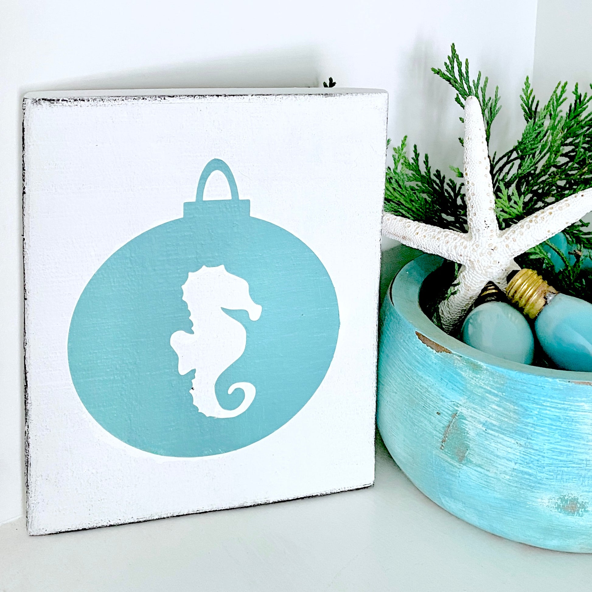 A square white wall sign with a distressed edge, featuring an aqua ornament design with a white seahorse silhouette. The sign is styled on a white surface next to a blue bowl containing a white starfish, vintage christmas bulbs and green foliage