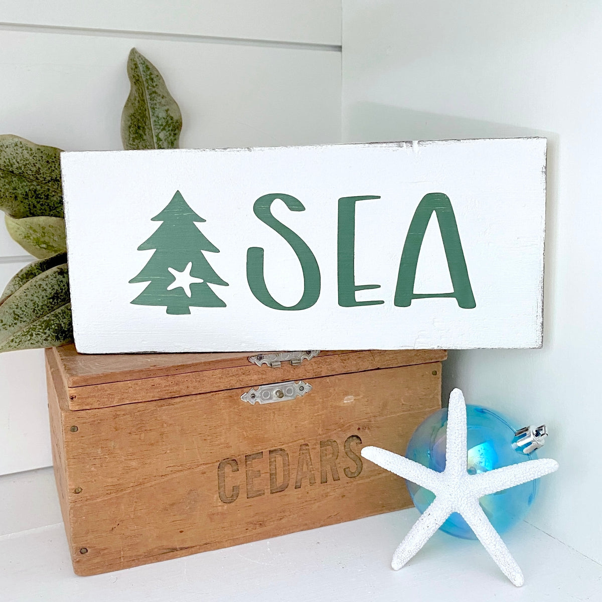 A white rectangular wooden sign with the word "SEA" in dark green and a green tree icon, styled on top of a wooden crate with a blue starfish ornament and green leaves nearby.