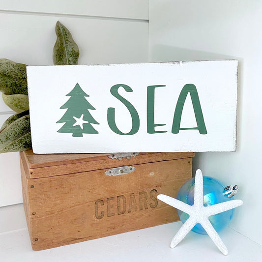 A white rectangular wooden sign with the word "SEA" in dark green and a green tree icon, styled on top of a wooden crate with a blue starfish ornament and green leaves nearby.