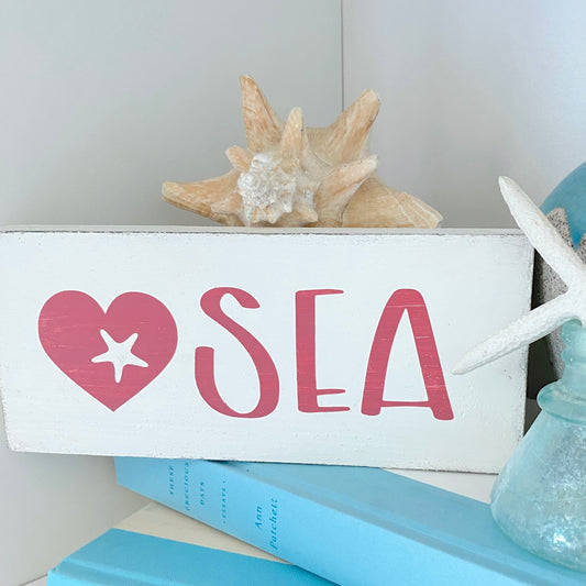 A white rectangular wooden sign with a pink heart containing a white starfish and the word 'SEA' in pink, styled with a seashell on top and blue books nearby.