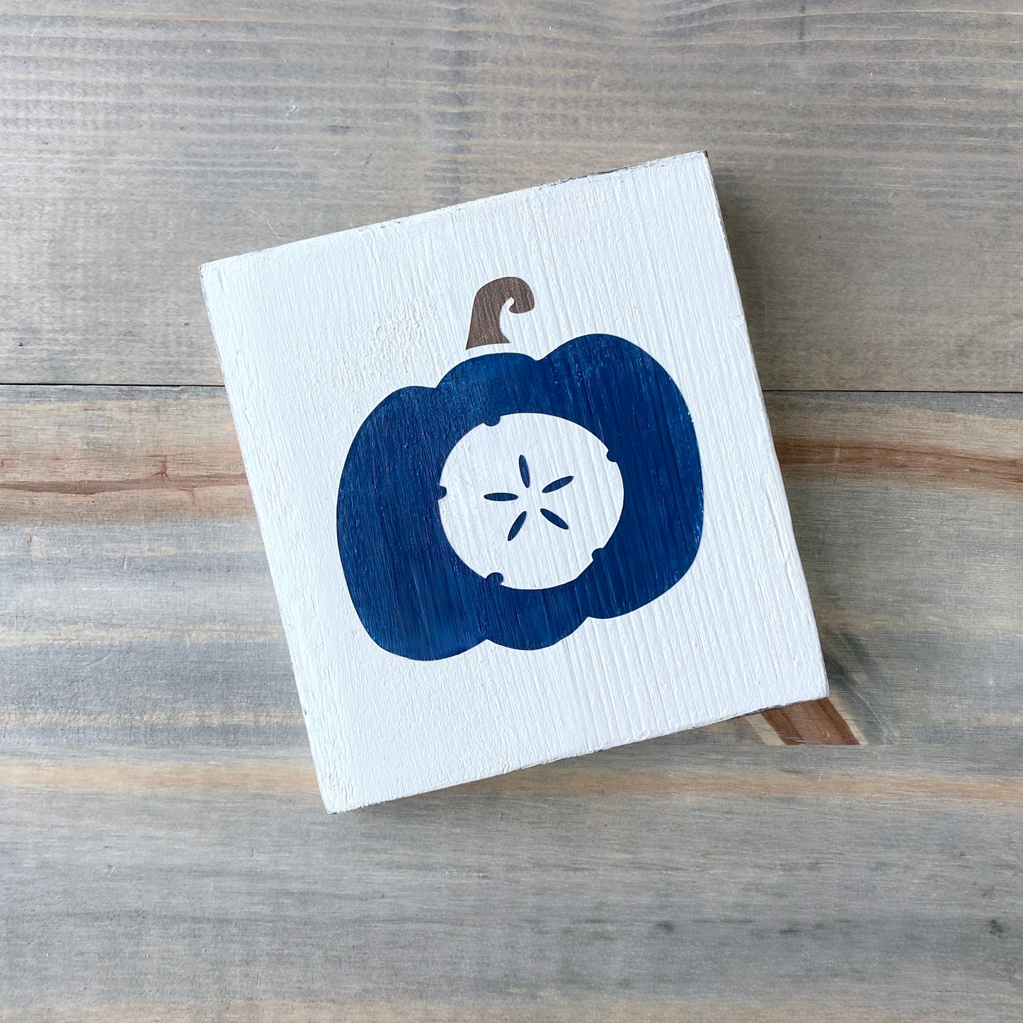 A square white wooden sign featuring a painted navy blue pumpkin with a white sand dollar motif in the center and a brown stem, displayed on a rustic gray wood plank surface.