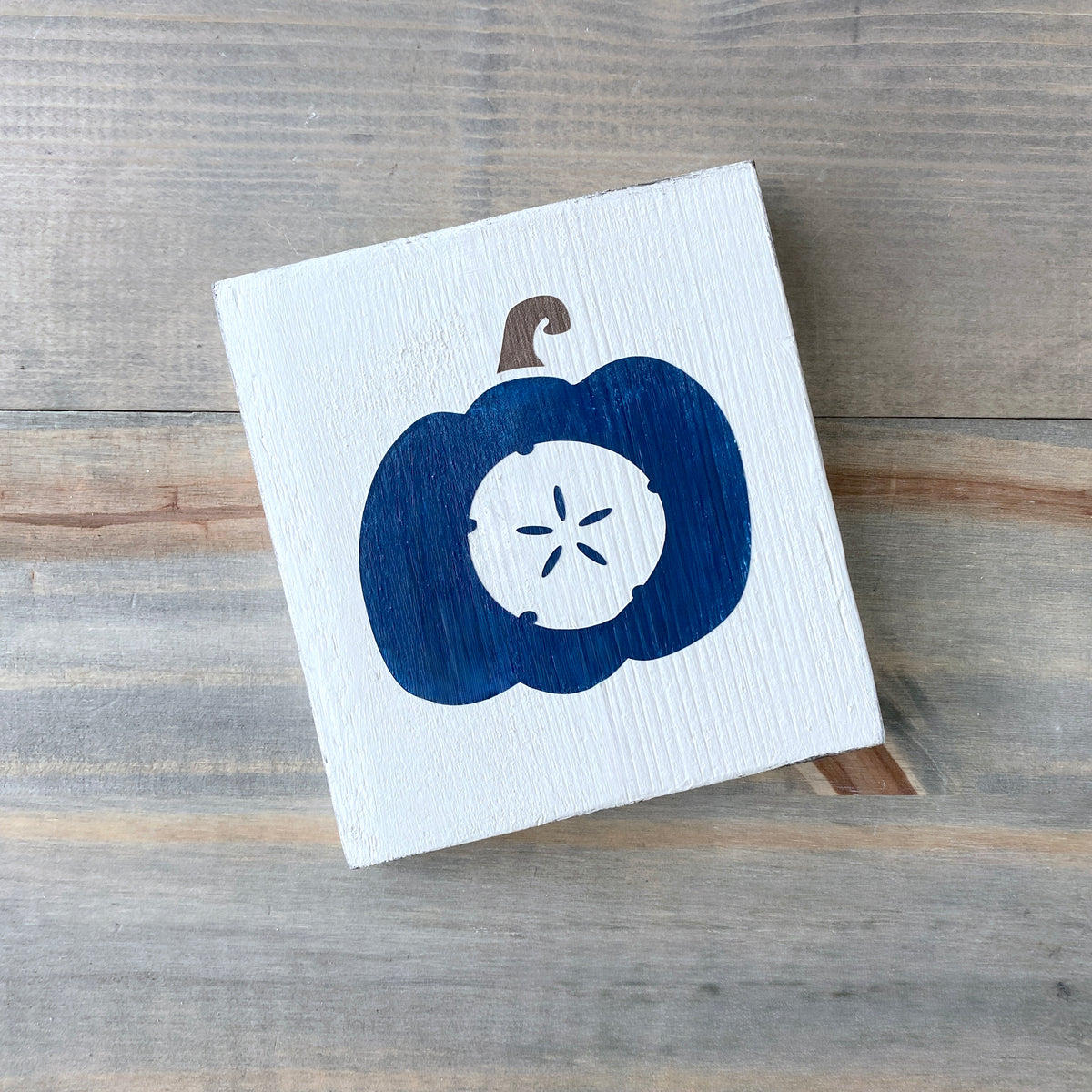 A square white wooden sign featuring a painted navy blue pumpkin with a white sand dollar motif in the center and a brown stem, displayed on a rustic gray wood plank surface.
