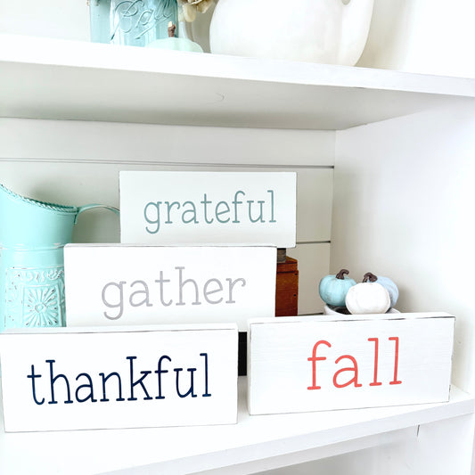 Decorative wooden blocks with 'grateful', 'gather', 'thankful', and 'fall' text on a shelf.