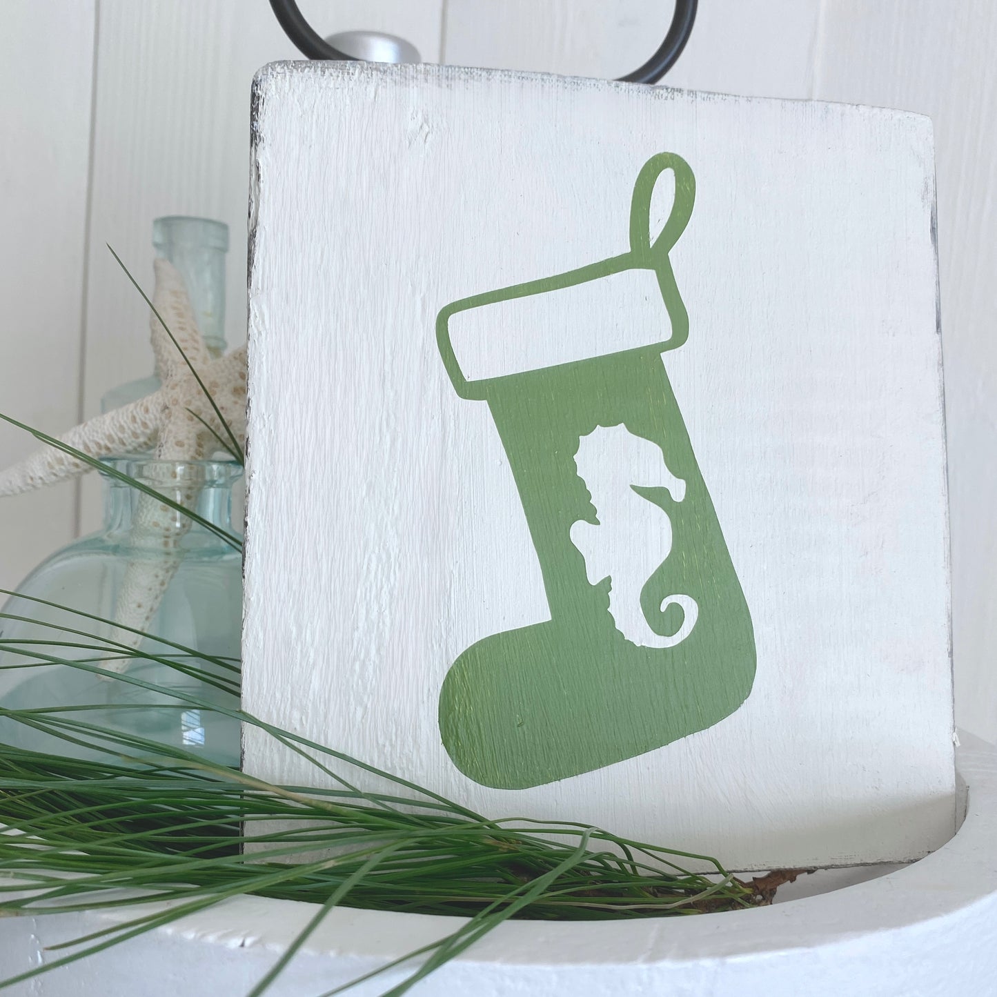 A square white wooden sign featuring a green Christmas stocking with a white seahorse silhouette, styled with green pine needles and a glass bottle in the background.