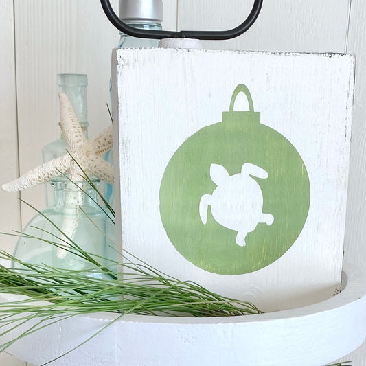 A rustic white wooden sign featuring a green circular ornament design with a white turtle silhouette. The sign is styled on a white pedestal with a starfish, a glass bottle, and green pine sprig against a white paneled background.