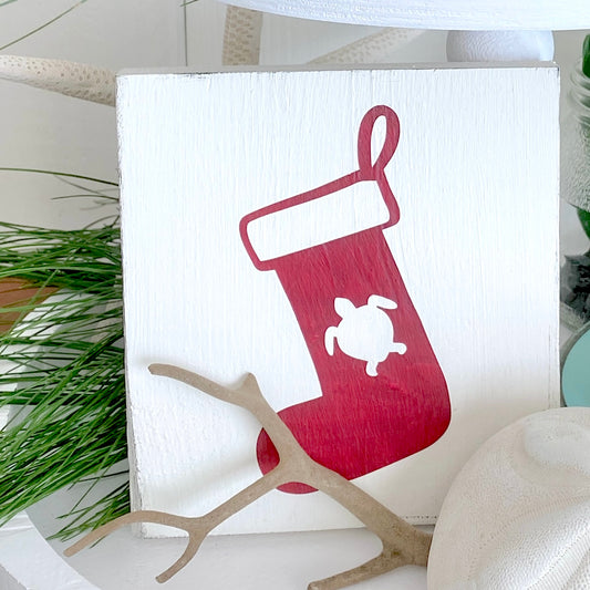 A square white wooden sign featuring a red Christmas stocking with a white turtle silhouette, styled with a small piece of driftwood and a pine sprig in a coastal home decor setting.