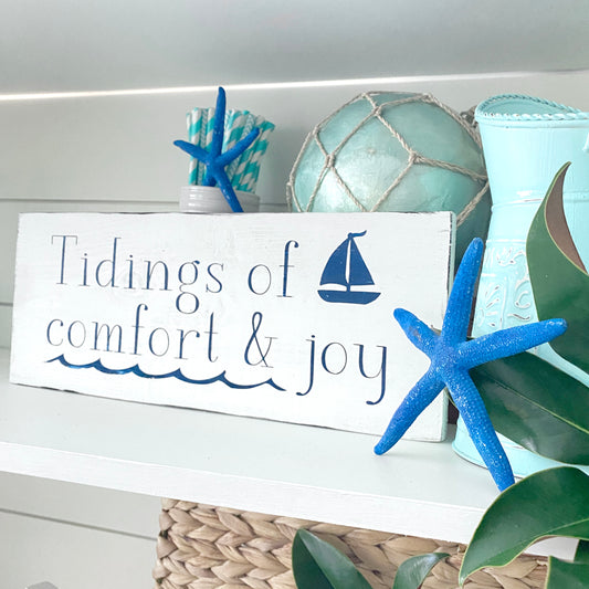 A white rustic wooden sign with the phrase 'Tidings of comfort & joy' in navy blue lettering and a small sailboat graphic, styled on a shelf with blue starfish, a mint fishing float and magnolia leaves.