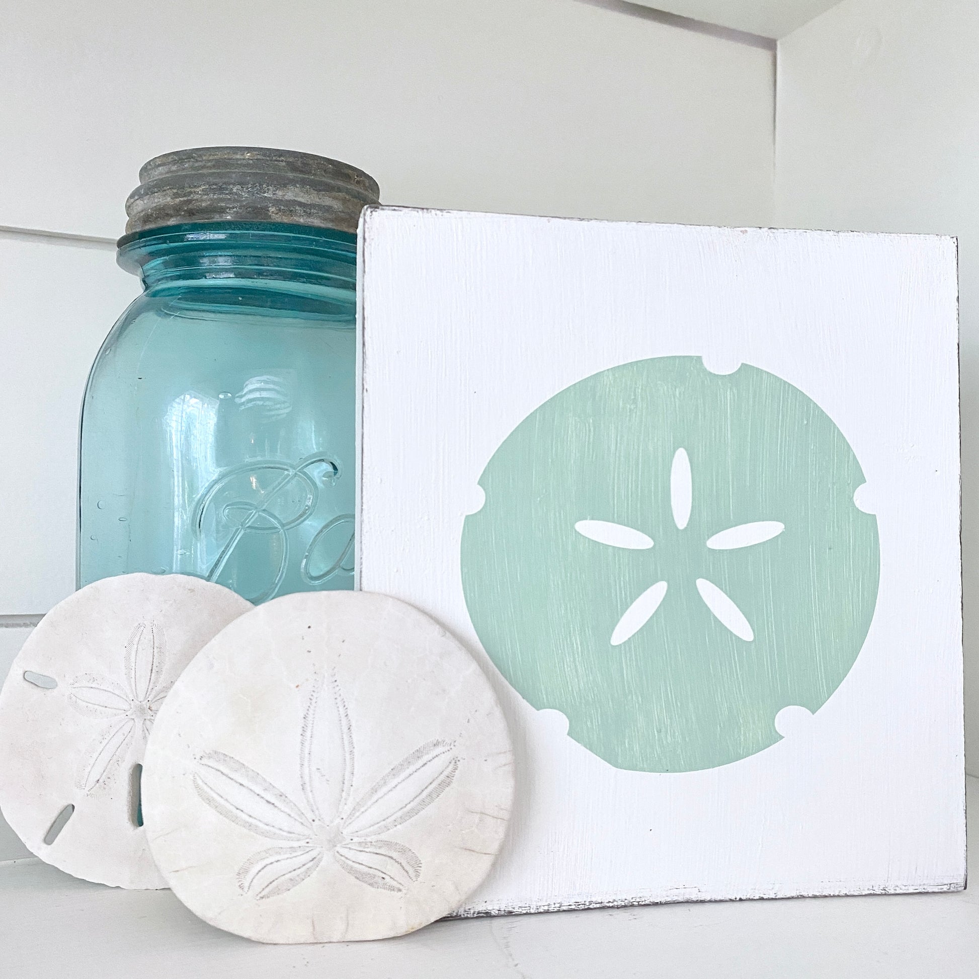 A square white painted canvas featuring a mint green sand dollar design, styled on a white surface next to a vintage-style blue glass jar and two real sand dollars.