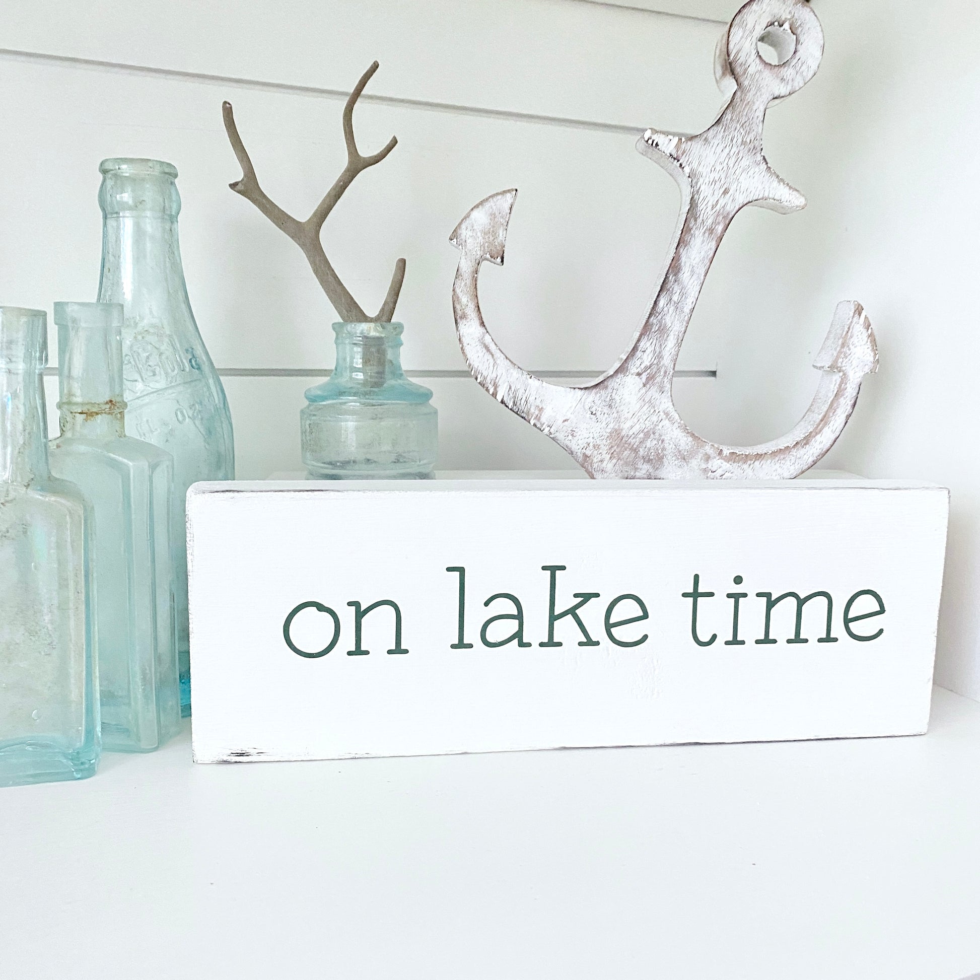 A white rectangular decorative sign with the words 'on lake time' in dark green lowercase letters, styled on a white surface with a distressed white anchor, pale blue glass bottles, and a small antler holder in the background.