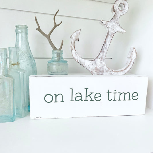 A white rectangular decorative sign with the words 'on lake time' in dark green lowercase letters, styled on a white surface with a distressed white anchor, pale blue glass bottles, and a small antler holder in the background.