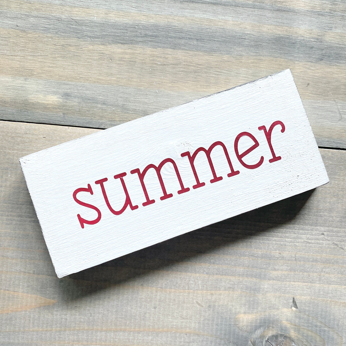 A rectangular white wooden sign with the word 'summer' in red lowercase, photographed on a rustic gray wood plank surface.