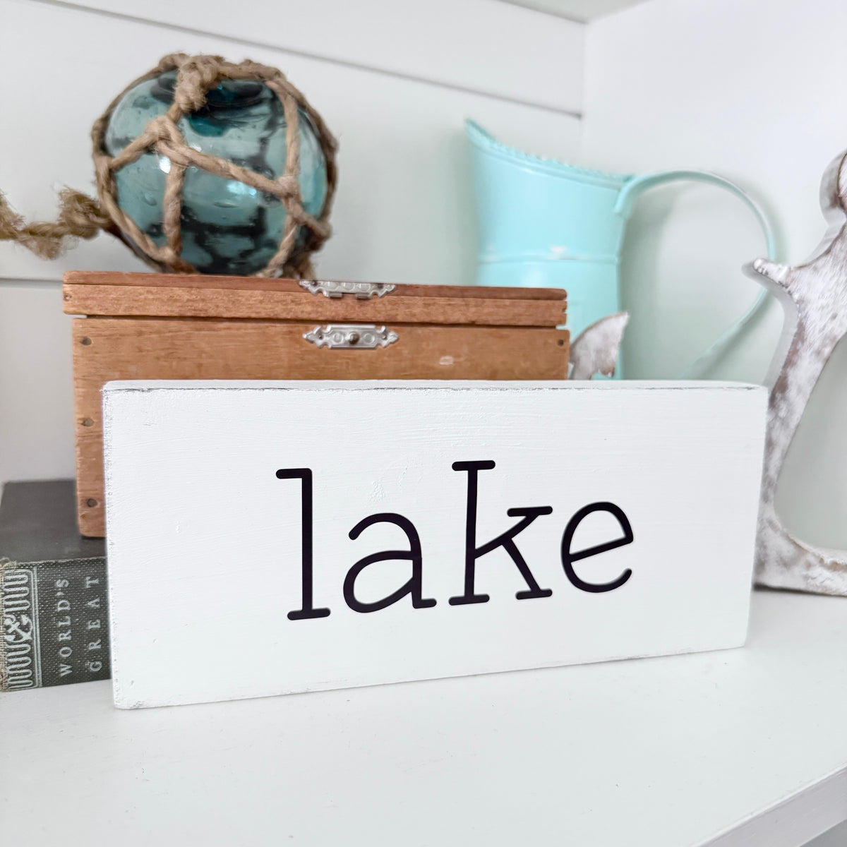 A white rectangular wooden sign with the word 'lake' in black lowercase styled on a white shelf with a light wood box, a blue-green rope ball, a mint pitcher, and other home decor in the background.