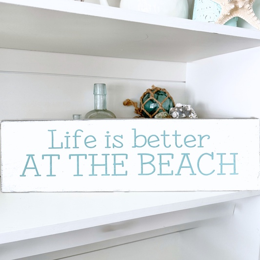 A white rectangular wooden sign with the phrase 'Life is better AT THE BEACH' in aqua blue lettering, displayed on a white shelf with decorative beach-themed items.