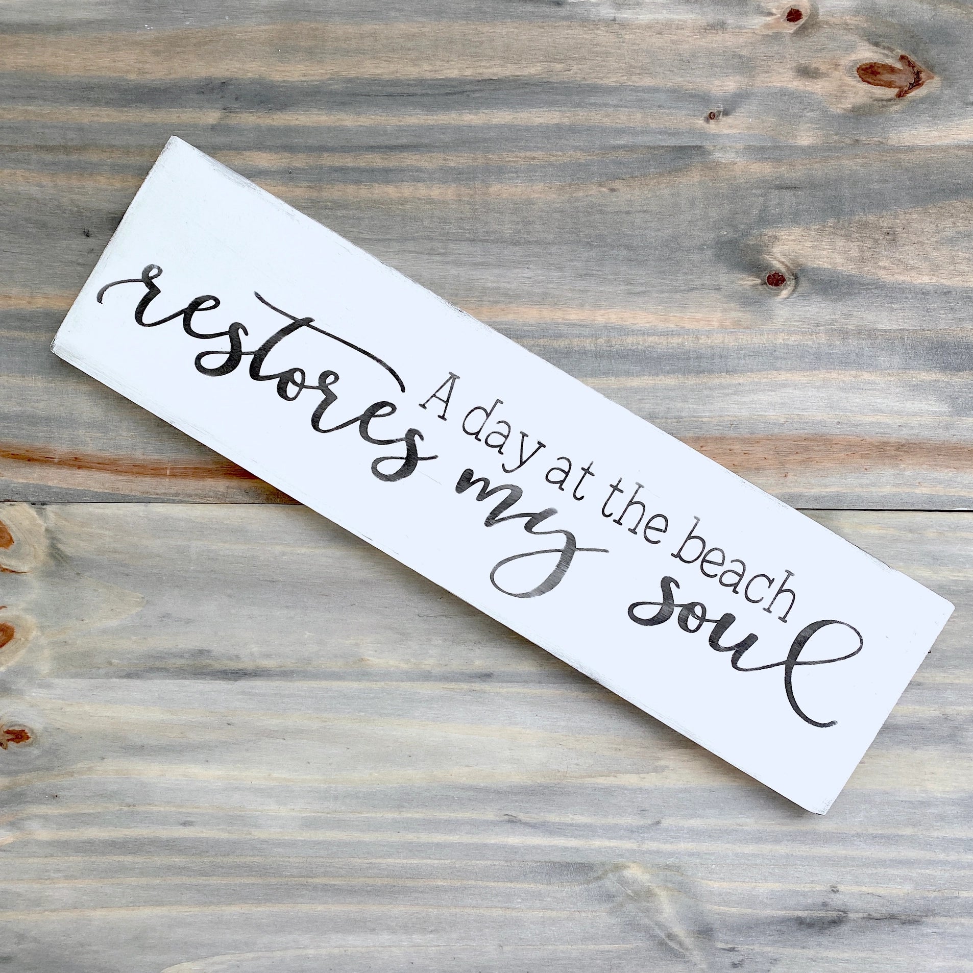 White sign with black text 'A day at the beach restores my soul' on a wooden surface