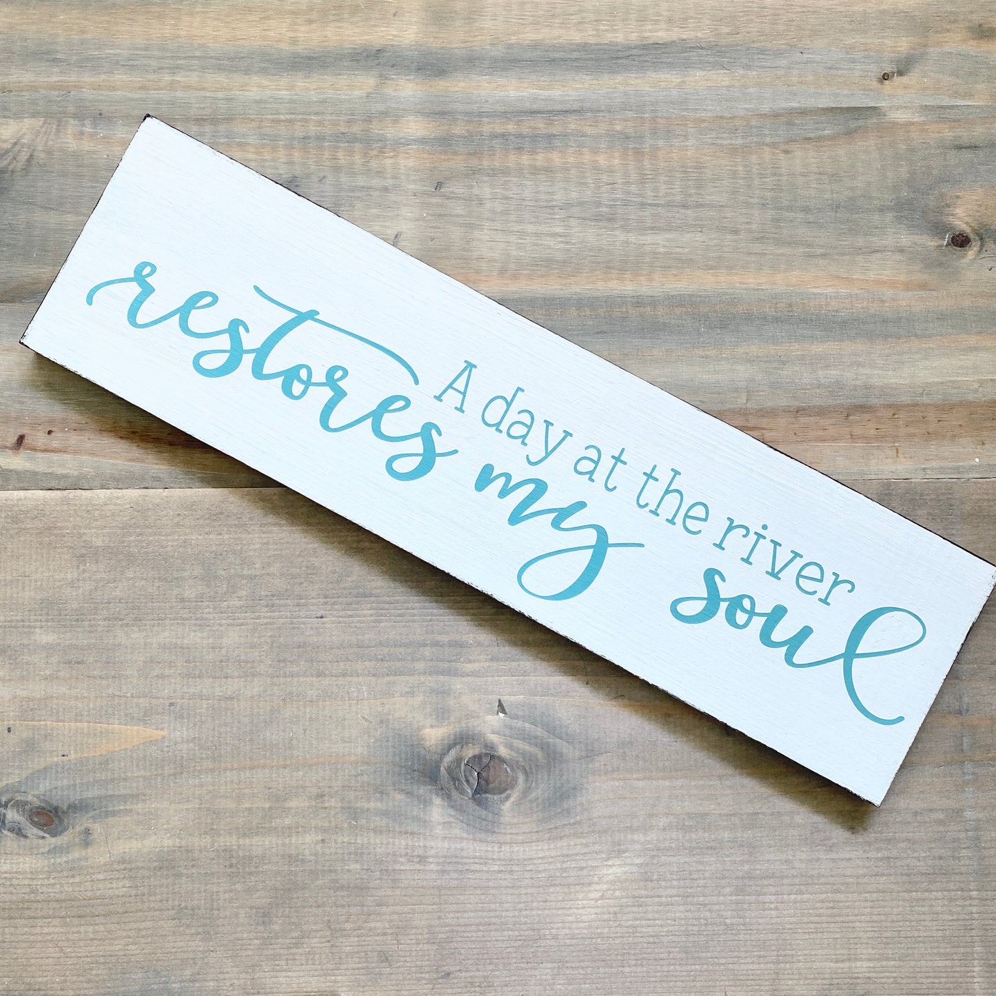 Wooden sign with text 'A day at the river restores my soul' on a wooden surface