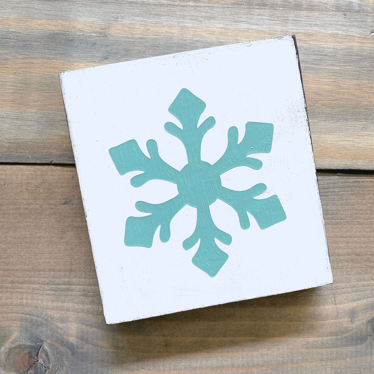 A square white wooden sign with an aqua snowflake design, photographed from above on a rustic wooden surface.