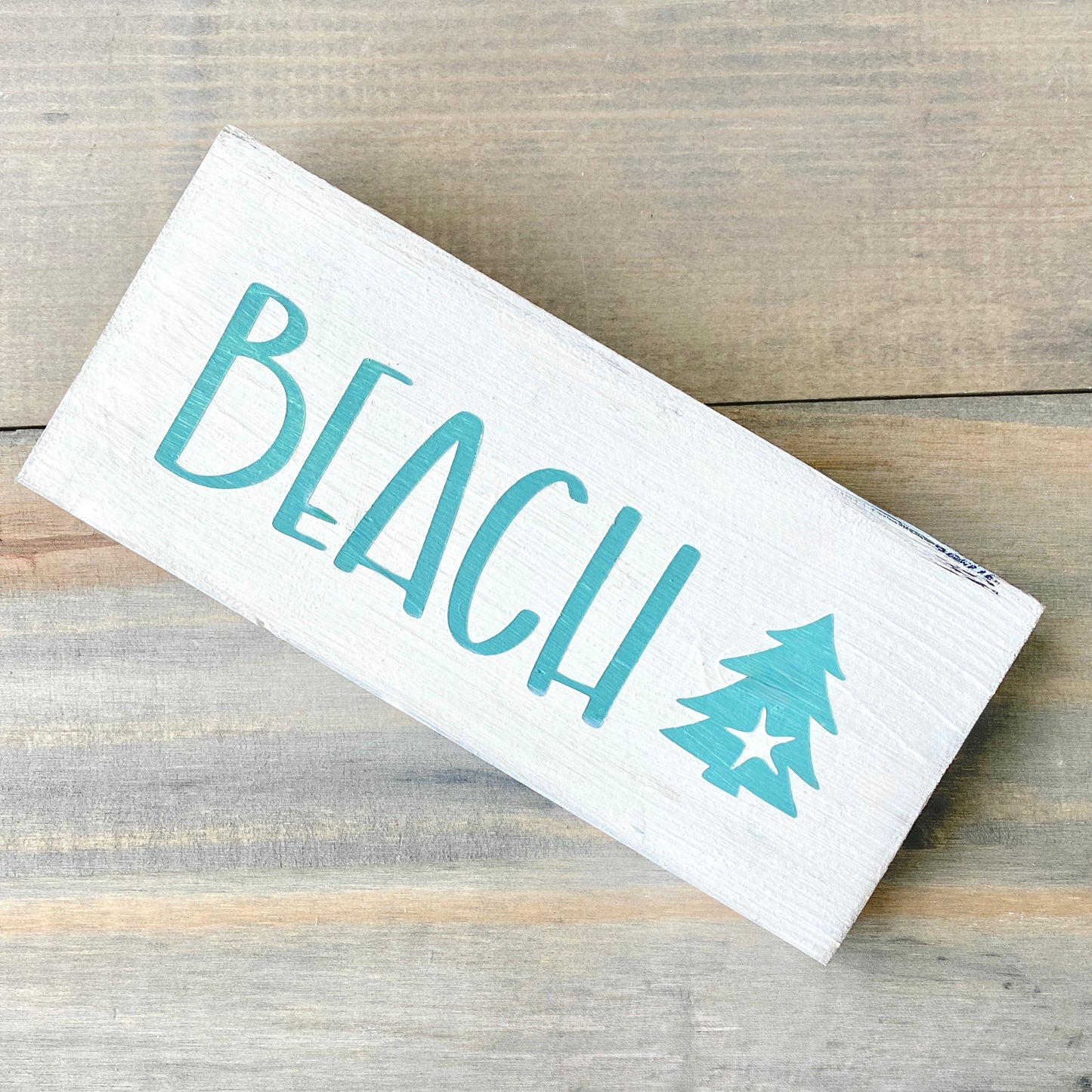 White wood sign with 'BEACH' and a christmas tree design on a wooden surface