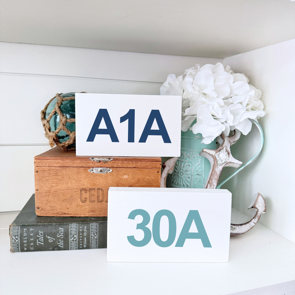 Two white rectangular sign plaques with navy and teal lettering reading 'A1A' and '30A', styled on a white shelf with a wooden chest, books, white flowers, and a turquoise vase.