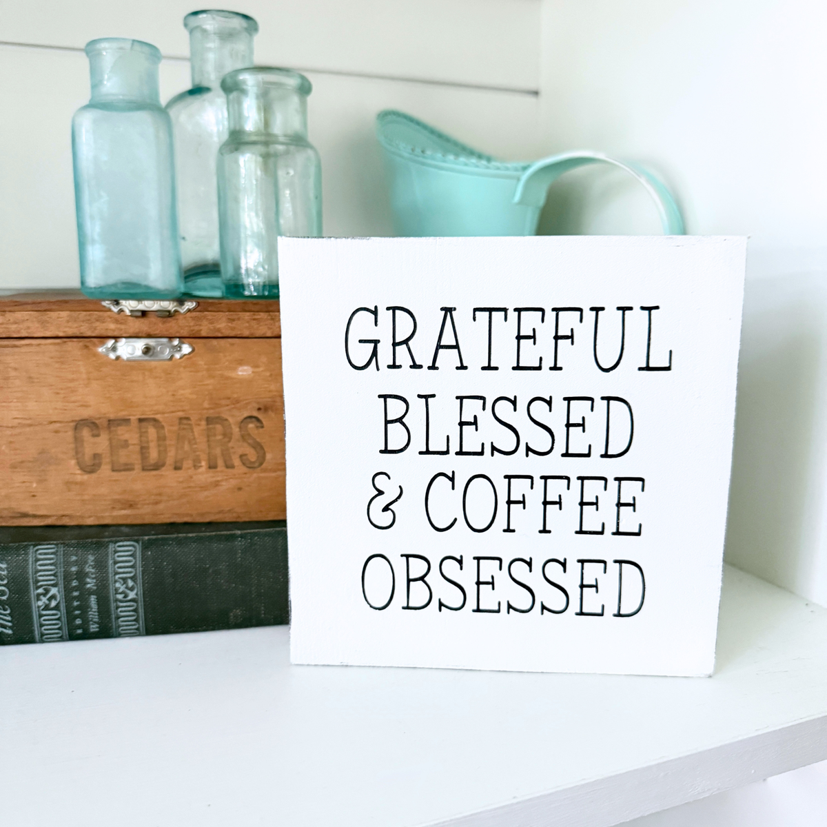 A white square sign with black text reading 'GRATEFUL BLESSED & COFFEE OBSESSED' displayed on a white shelf next to decorative glass bottles and a wooden box.