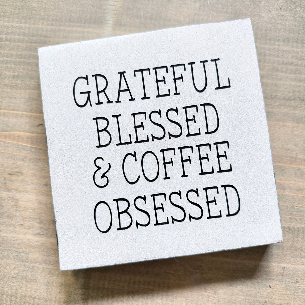 A square white canvas wall sign with the phrase 'GRATEFUL BLESSED & COFFEE OBSESSED' in black lettering, displayed on a light brown wooden surface.