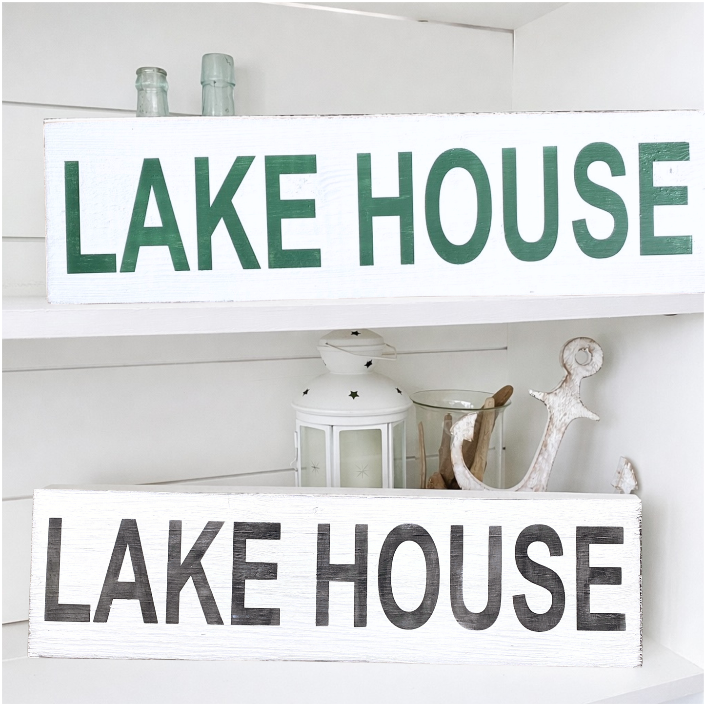 Two rustic white wooden signs with 'LAKE HOUSE' lettering, displayed on white shelves. The top sign has green letters, and the bottom sign has black distressed letters.