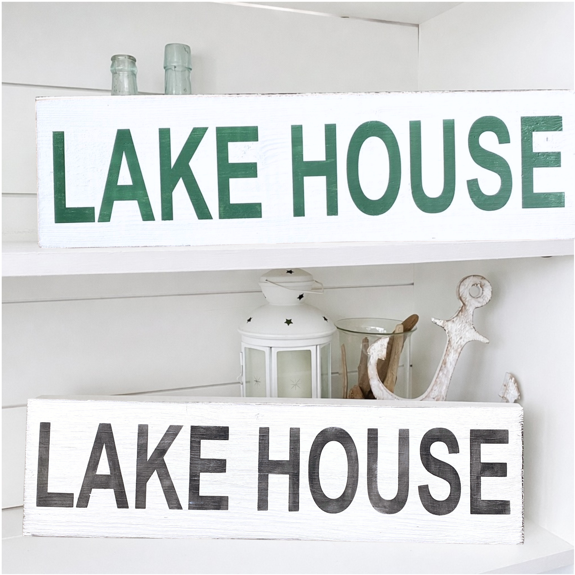 Two rustic white wooden signs with 'LAKE HOUSE' lettering, displayed on white shelves. The top sign has green letters, and the bottom sign has black distressed letters.