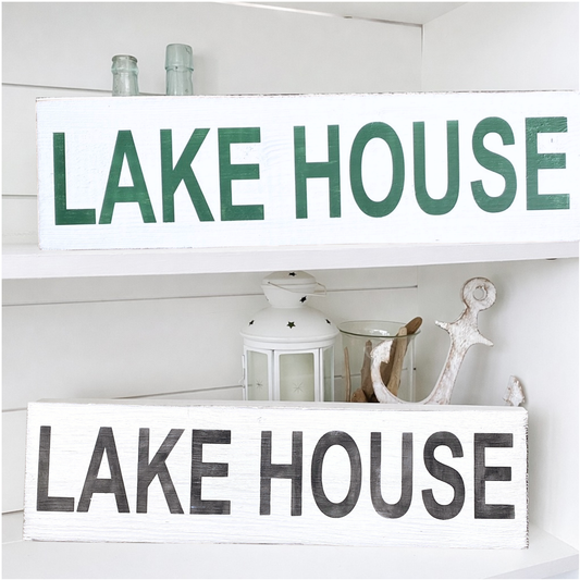 Two rustic white wooden signs with 'LAKE HOUSE' lettering, displayed on white shelves. The top sign has green letters, and the bottom sign has black distressed letters.