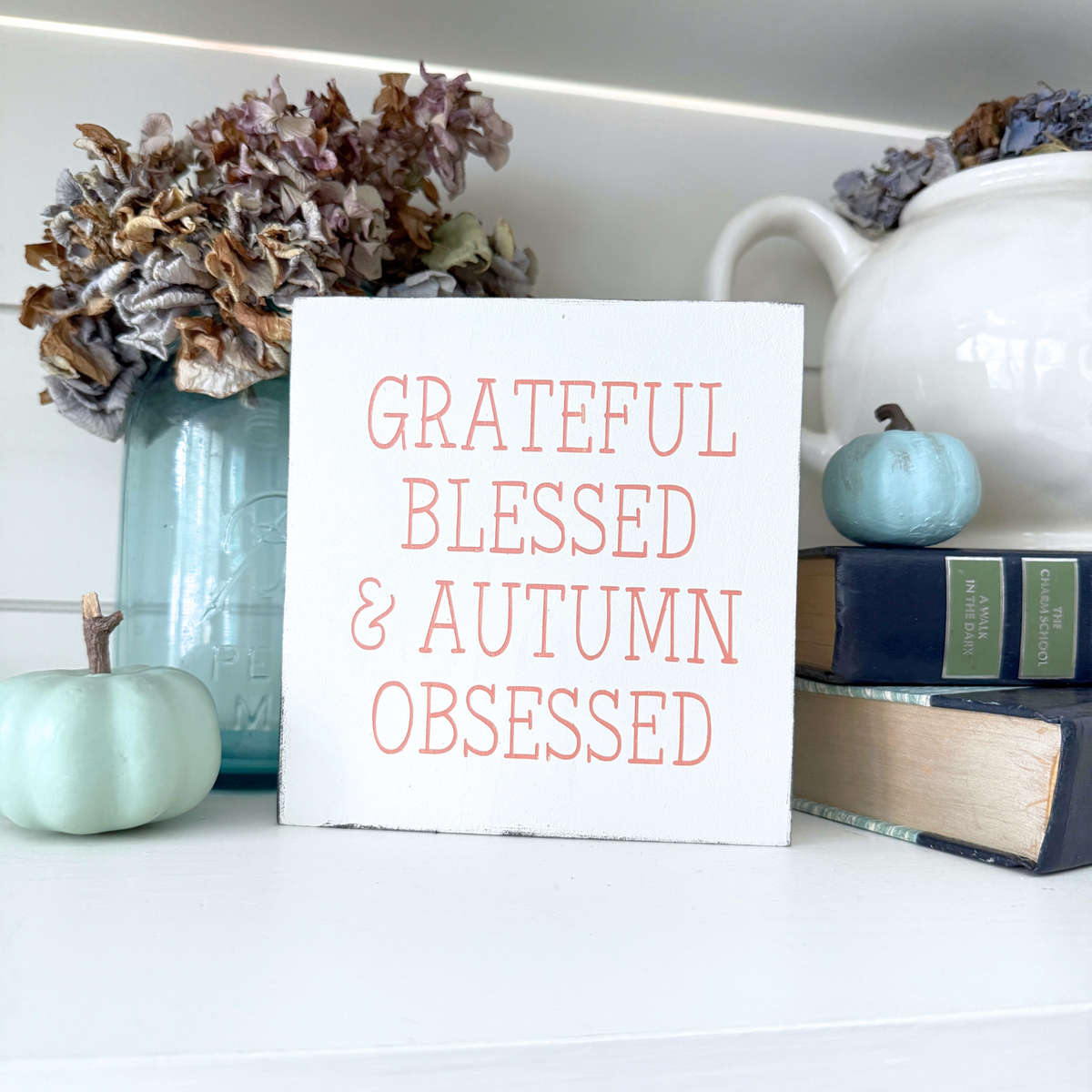 A white rectangular decorative sign with the phrase “GRATEFUL BLESSED & AUTUMN OBSESSED” in pale orange lettering, styled on a white shelf with dried flowers in a blue mason jar, a small mint pumpkin, books, and a white teapot.