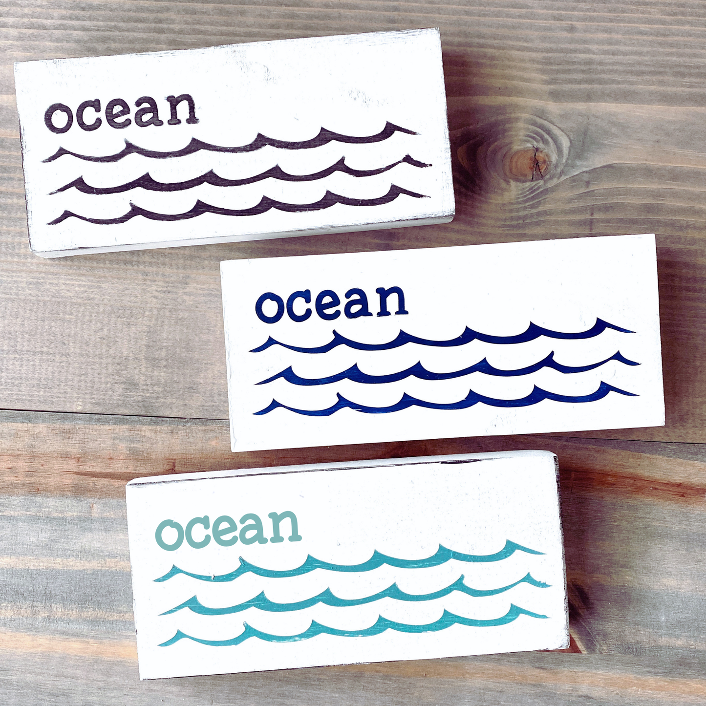 Three rectangular white wooden coasters with the word 'ocean' and wave illustrations, shown on a rustic wood surface. The coasters are white with dark purple, navy blue, and teal lettering and wave patterns.
