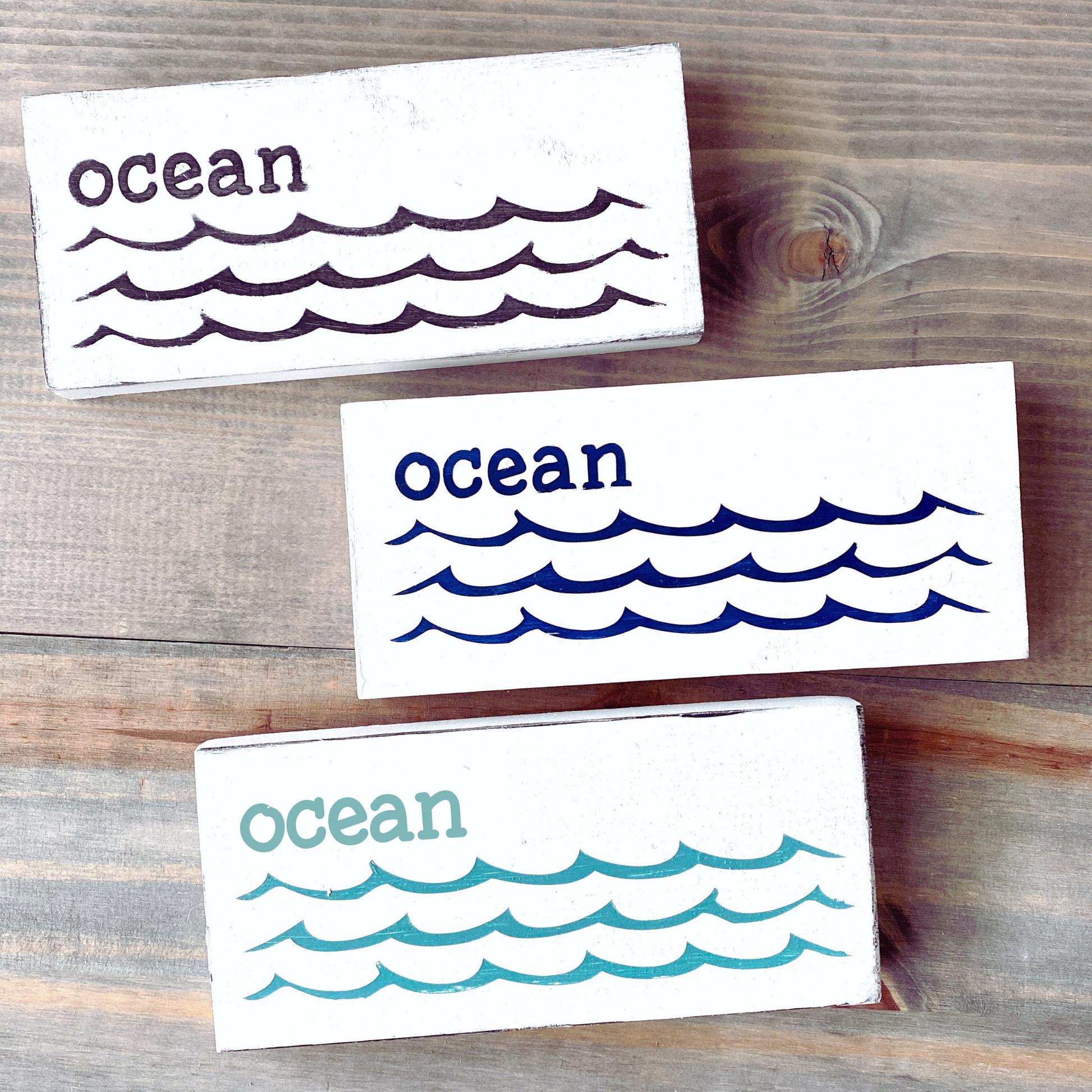 Three rectangular white wooden coasters with the word 'ocean' and wave illustrations, shown on a rustic wood surface. The coasters are white with dark purple, navy blue, and teal lettering and wave patterns.