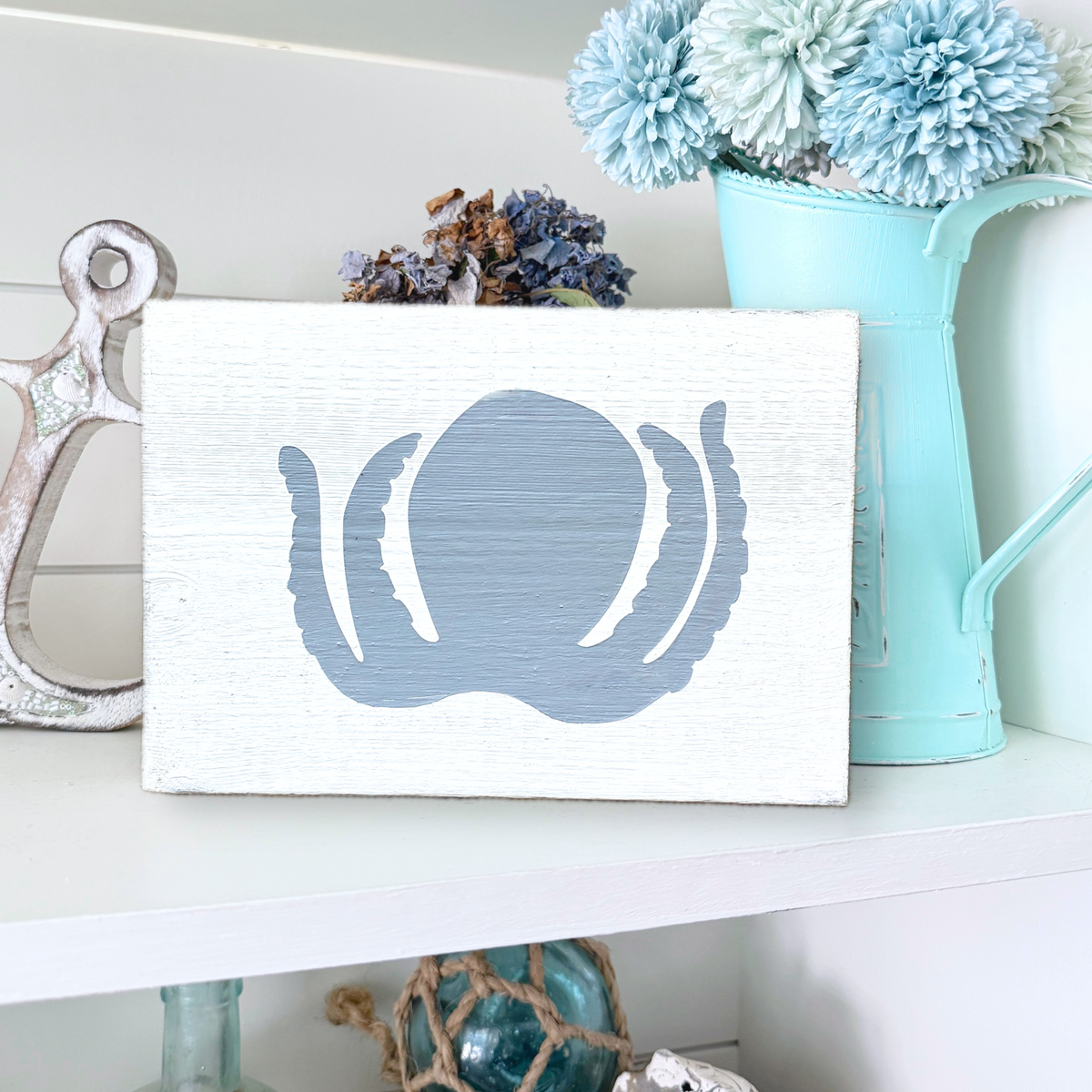 A rectangular white wooden sign with a distressed finish, featuring a painted blue octopus silhouette on the front. The sign is styled on a white shelf next to a silver anchor, dried flowers, and a light blue pitcher filled with blue pom-pom flowers.