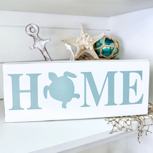 A white rectangular decorative sign with the word HOME in large sage green letters, where the O is replaced by a sea turtle silhouette. The sign is styled on a white shelf with nautical props, including a starfish, a small anchor, and a blue-green fishing float