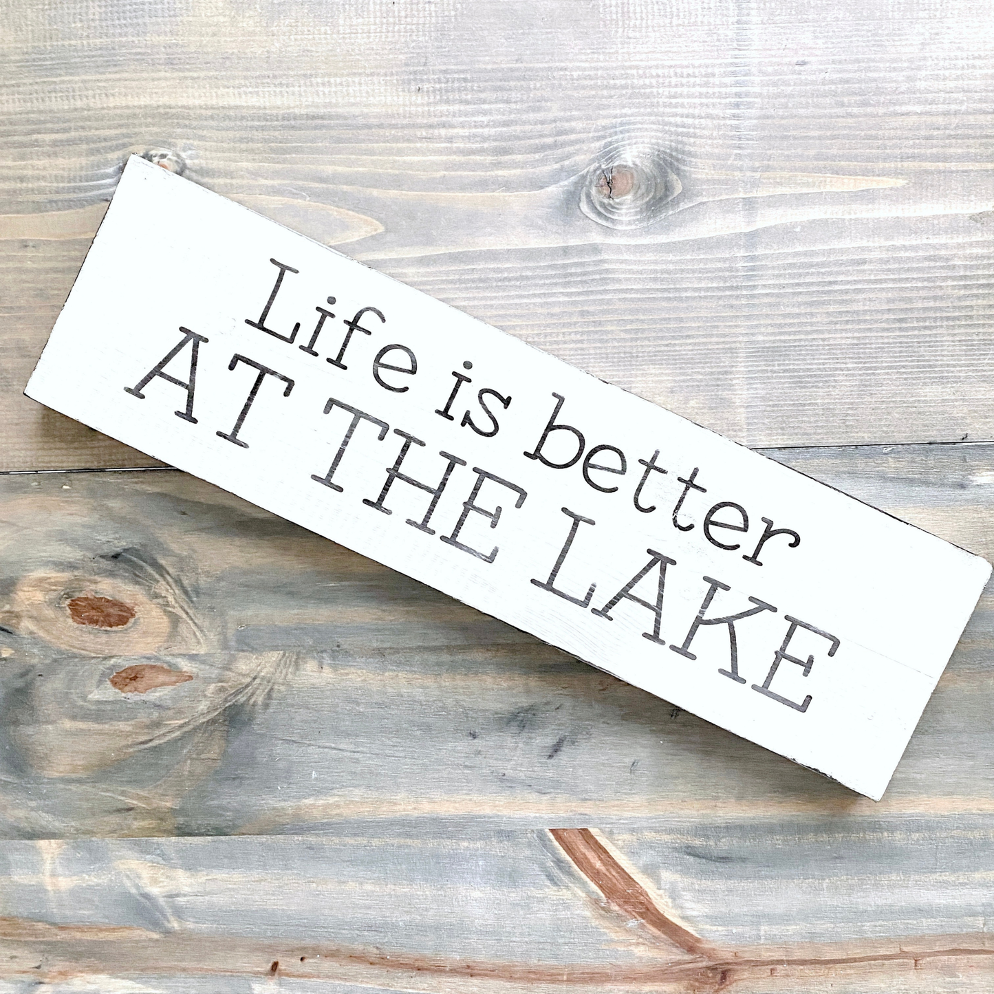 A rustic white wooden sign with the phrase 'Life is better AT THE LAKE' in black lettering, displayed diagonally on a weathered gray wood plank background.