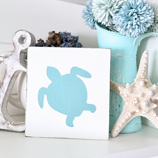 A square white decorative sign with an aqua sea turtle silhouette, styled on a shelf with a white anchor, a beige starfish, a turquoise bucket of flowers, and dried blue-purple flowers.
