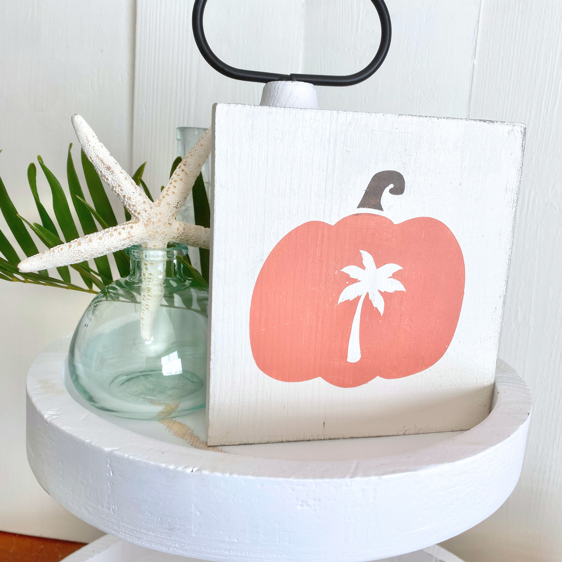A square white wooden sign featuring a coral-pink pumpkin with a white palm tree motif, styled on a white pedestal stand next to a glass vase with a starfish and green leaves.