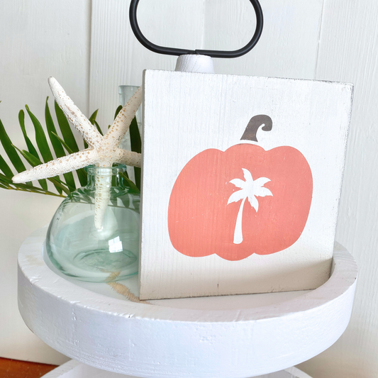 A square white wooden sign featuring a coral-pink pumpkin with a white palm tree motif, styled on a white pedestal stand next to a glass vase with a starfish and green leaves.