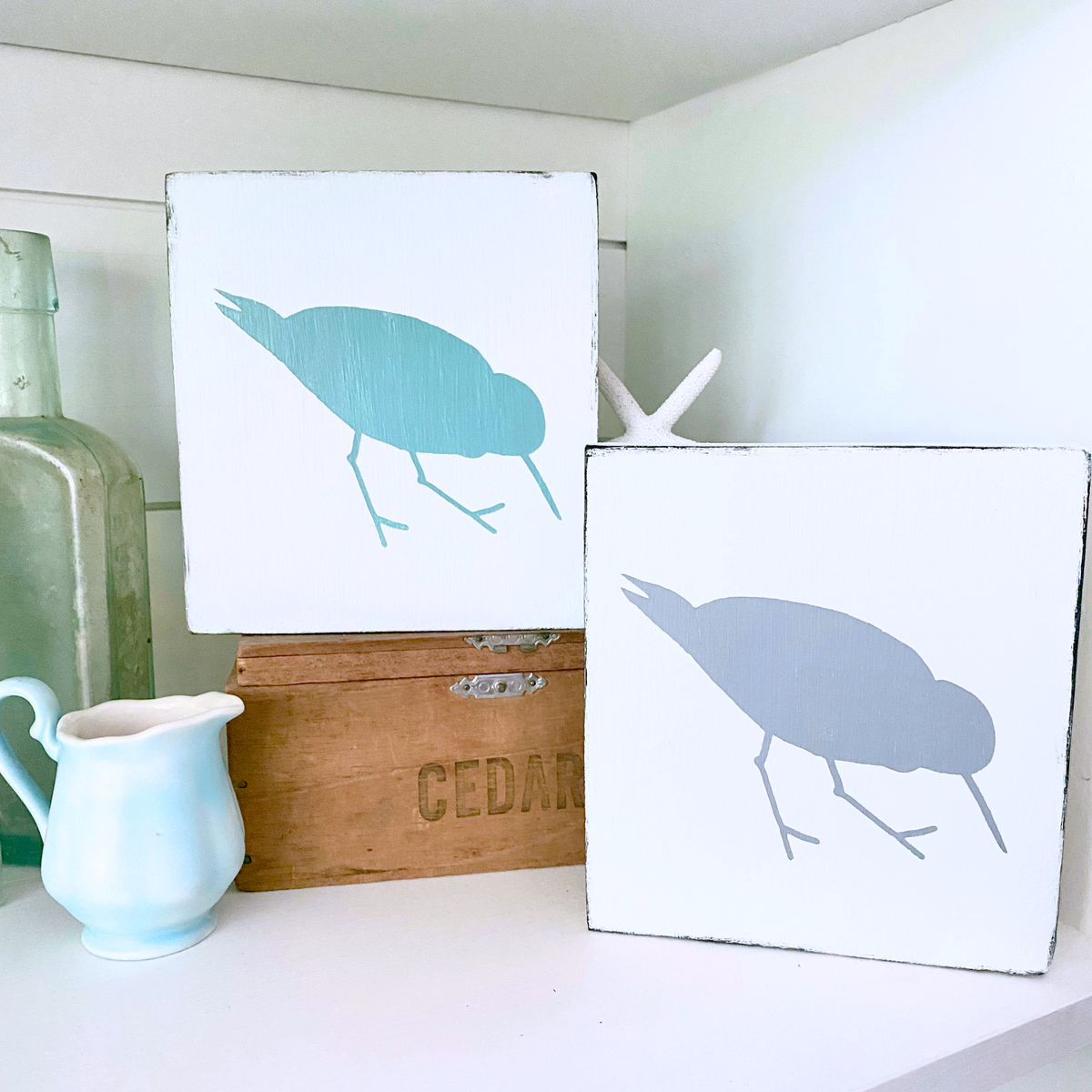Two square white decorative wall art signs with shorebird, sandpiper silhouettes, displayed on a shelf beside a small white pitcher, a wooden box, and a green glass bottle.