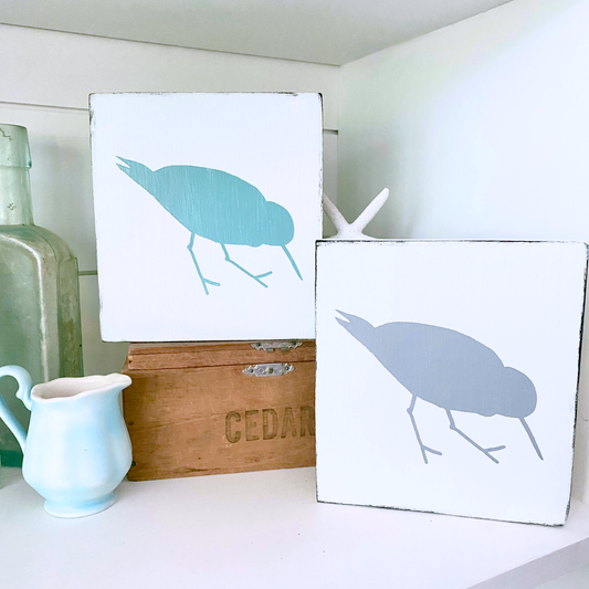 Two square white decorative wall art signs with shorebird, sandpiper silhouettes, displayed on a shelf beside a small white pitcher, a wooden box, and a green glass bottle.