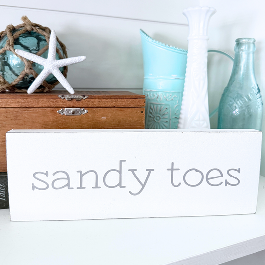 A white rectangular wooden sign with the words 'sandy toes' in gray lowercase font displayed on a white shelf in a styled home decor setting with a wooden box, starfish, and decorative bottles.