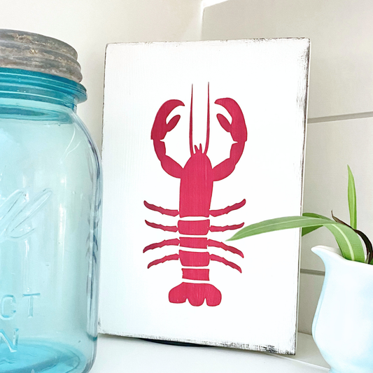 A rectangular white sign with a bright red lobster illustration, styled on a shelf next to a blue glass mason jar and a small potted plant.
