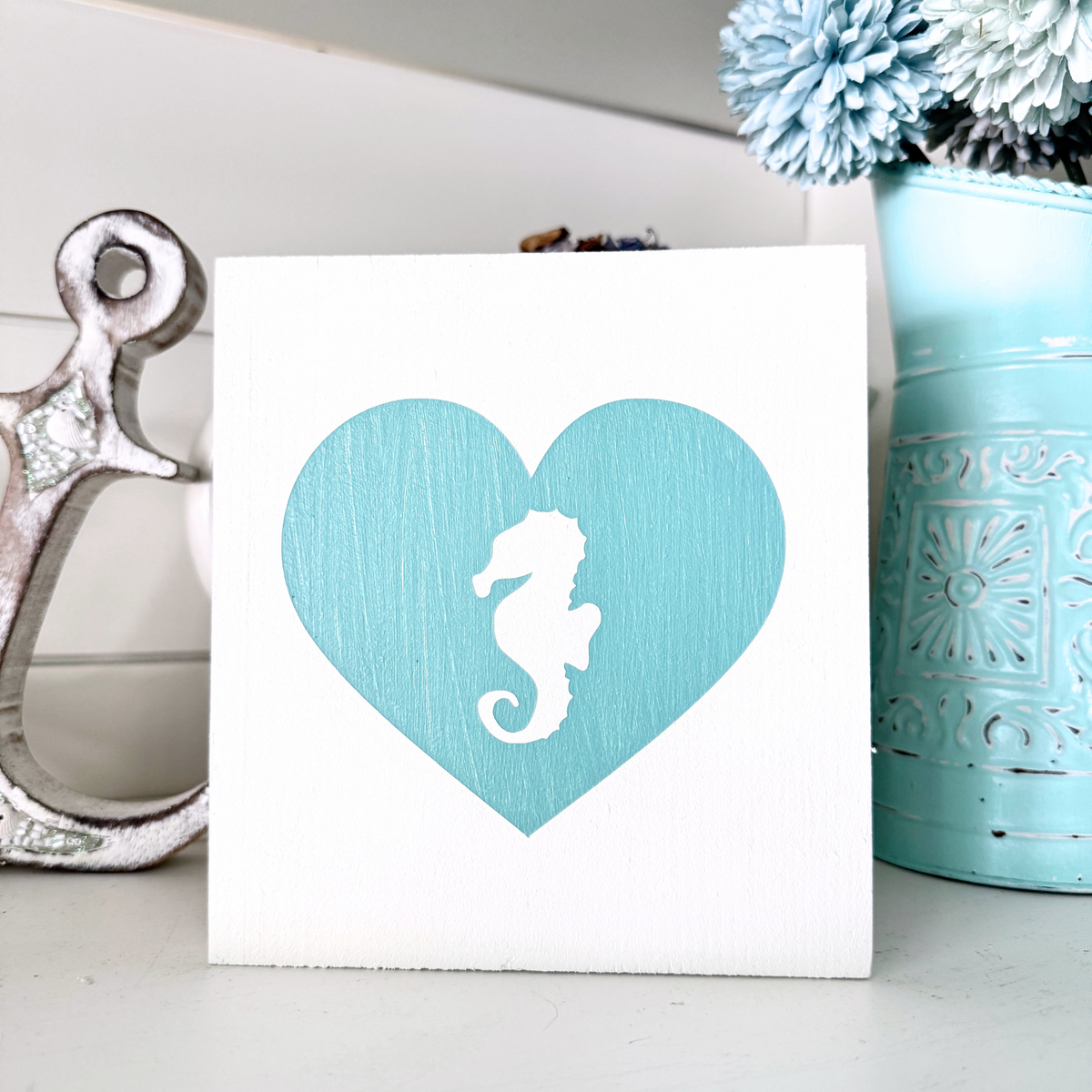 A square white wall art plaque featuring a large textured aqua heart with a white seahorse silhouette in the center, styled on a tabletop with a wood anchor and a aqua decorative vase in the background.