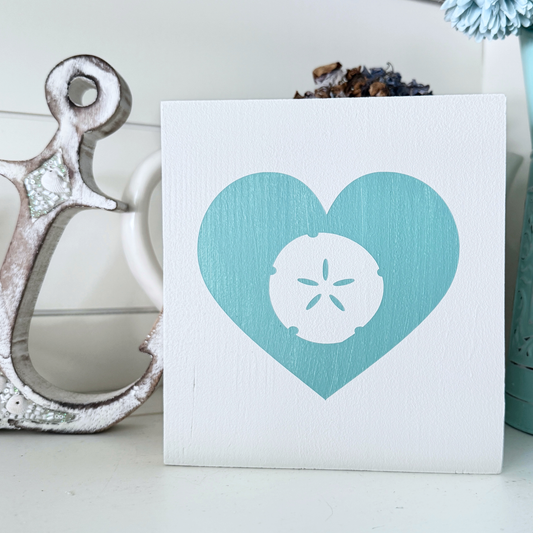 A square white wooden sign featuring a aqua heart with a white sand dollar design in the center, styled on a shelf next to a distressed wood anchor.