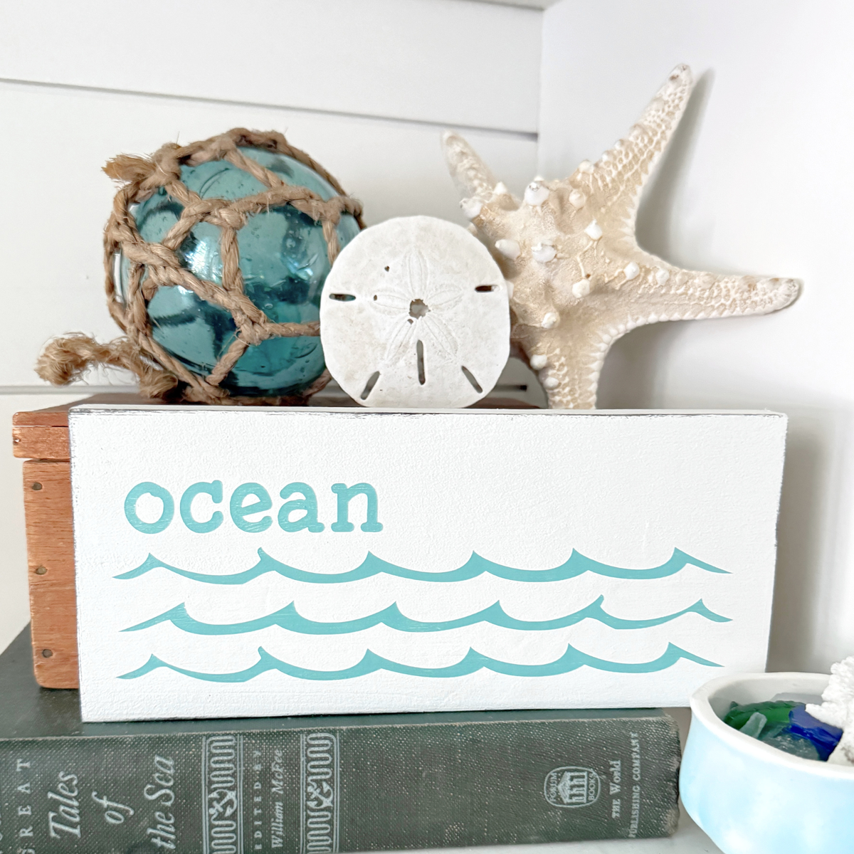 A white decorative block sign with the word 'ocean' and teal wave lines, styled on top of books with a blue glass orb in net, a sand dollar, and a starfish.