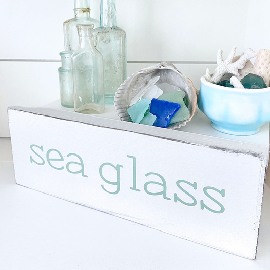 A white rectangular wooden sign with the words 'sea glass' in a light mint font, styled on a white surface with decorative glass bottles, a shell of sea glass, and a blue bowl of coral and sea glass