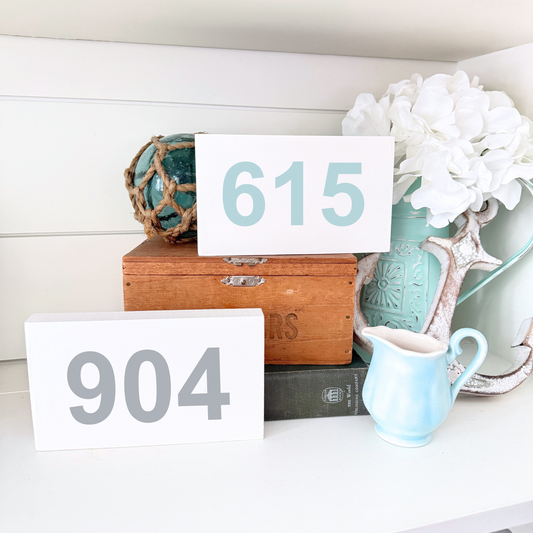 Two white rectangular house number signs, one with gray numerals reading 904 and the other with light blue numerals reading 615, displayed on a shelf with decorative props including a wooden box, white flowers, a turquoise pitcher, and a blue-green nautical ornament.